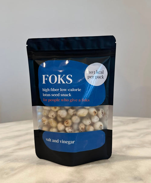 A stand-up pouch of FOKS lotus seed snack, salt and vinegar flavor, standing on a light marble-like surface against a plain light gray wall. The package is black with blue label areas and a clear window showing the beige lotus seeds inside.