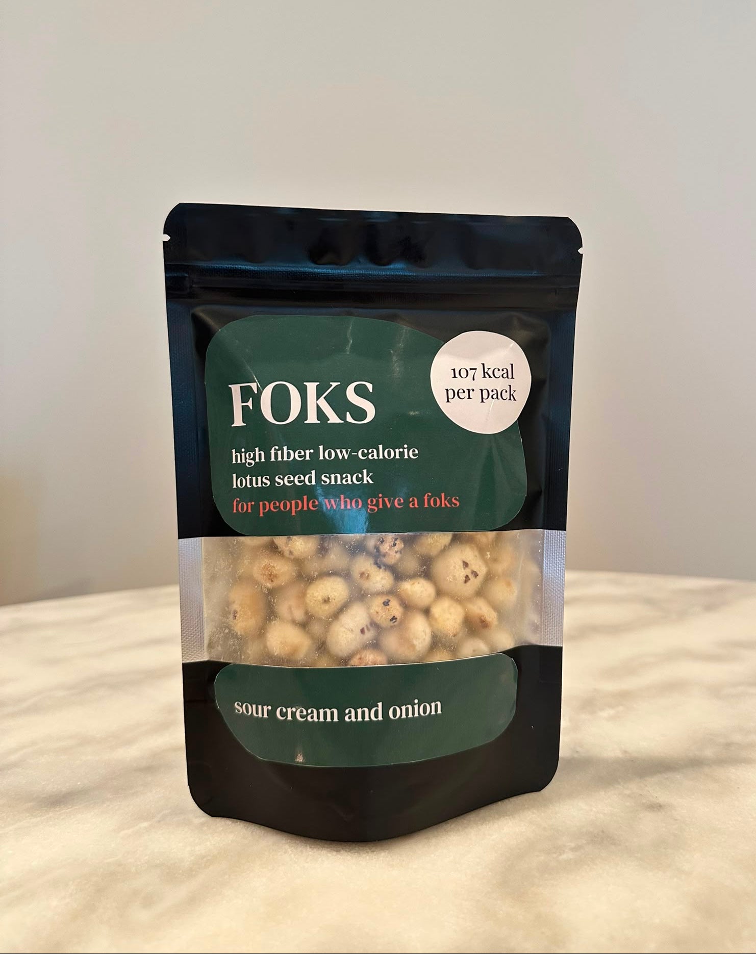 A stand-up pouch of Foks high fiber low-calorie lotus seed snack, featuring a dark green and black design with a clear window showing the light tan lotus seeds inside, set against a neutral beige background.